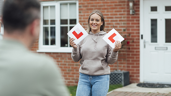 young lady walking out of the house towards a male ready to set off for a driving lesson holding two l plates
