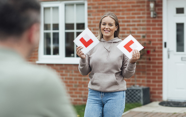 young lady holding l plates leaving house walking towards a male driving instructor