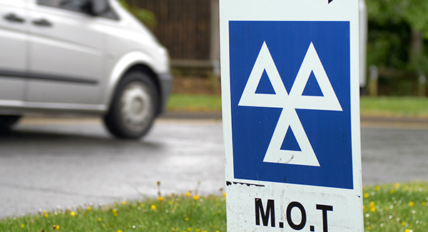 blue mot testing sign on grass by road with silver car driving past